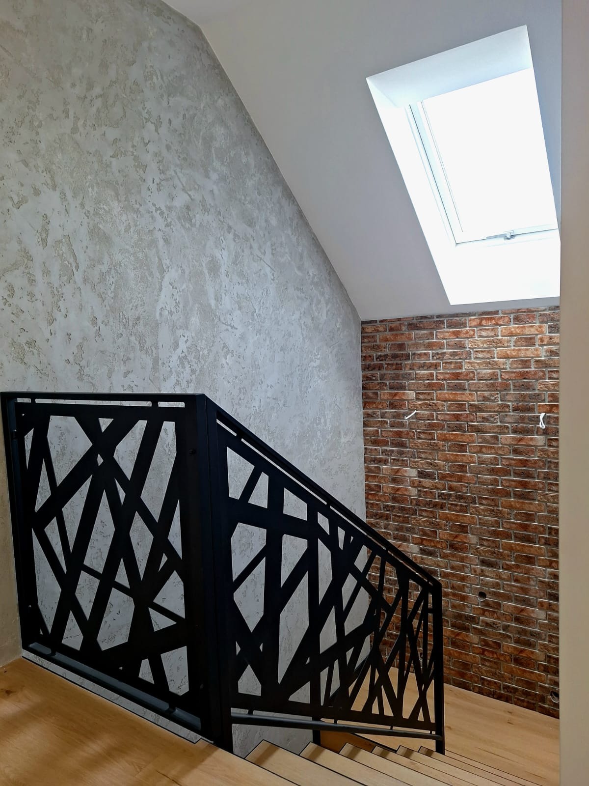 Schody z drewnianymi stopniami i nowoczesną, czarną, ażurową balustradą w Grudziądzu. Ściana pokryta dekoracyjnym tynkiem strukturalnym, fragment ściany wykończony czerwoną cegłą. Widoczne okno...