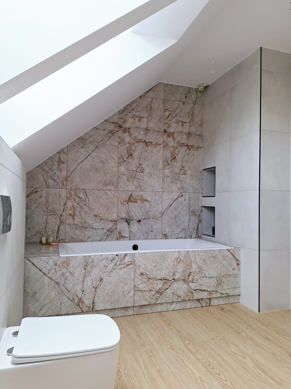 Wykończona łazienka na poddaszu z wanną obudowaną płytkami imitującymi marmur, drewnopodobną podłogą i oknem dachowym.