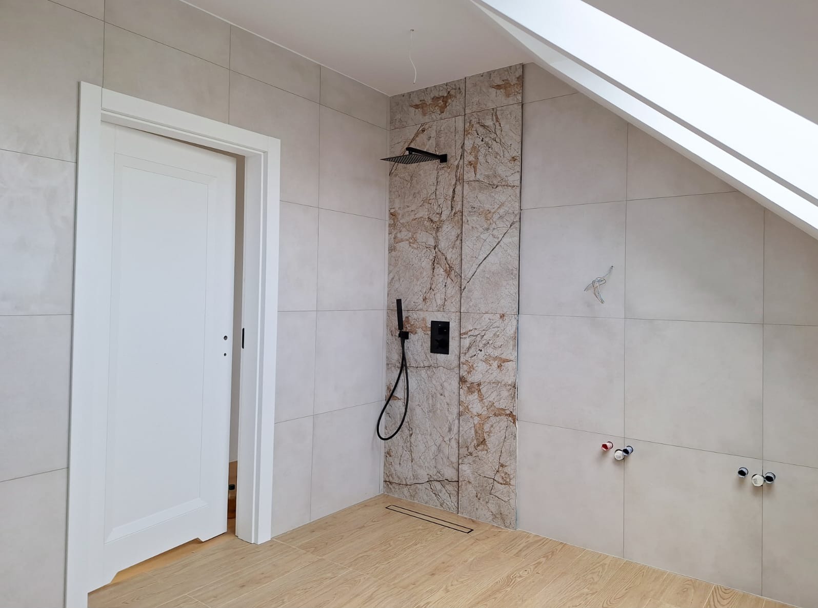 Nowoczesna łazienka na poddaszu z beżowymi płytkami, kabiną prysznicową typu walk-in z elementami marmuru i czarną armaturą, widoczne niedokończone podłączenia wodne.