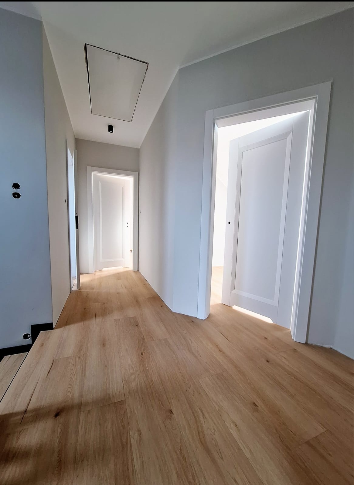 Nowoczesny korytarz z panelami podłogowymi w odcieniach drewna, świeżo pomalowane ściany na szaro, białe drzwi z minimalistycznymi ościeżnicami, widoczny właz rewizyjny w suficie.