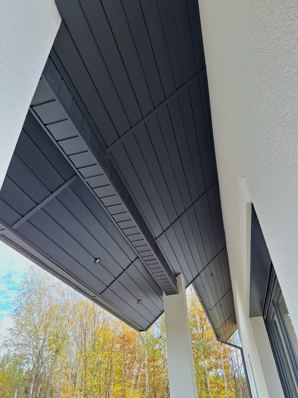 Nowoczesna zabudowa podbitki dachowej z oświetleniem punktowym, widok z dołu na tle jesiennych drzew.