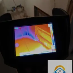 Kamera termowizyjna w trakcie pomiaru wilgotności ściany, widoczny ekran z kolorowym rozkładem temperatur i logo firmy w prawym dolnym rogu.