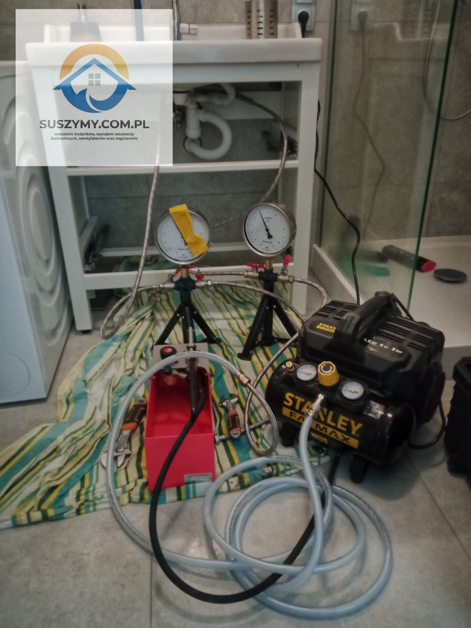 Sprzęt do pomiaru ciśnienia i testowania szczelności instalacji hydraulicznej, z manometrami, kompresorem Stanley Fatmax i wężami, rozłożony na podłodze w łazience obok pralki.