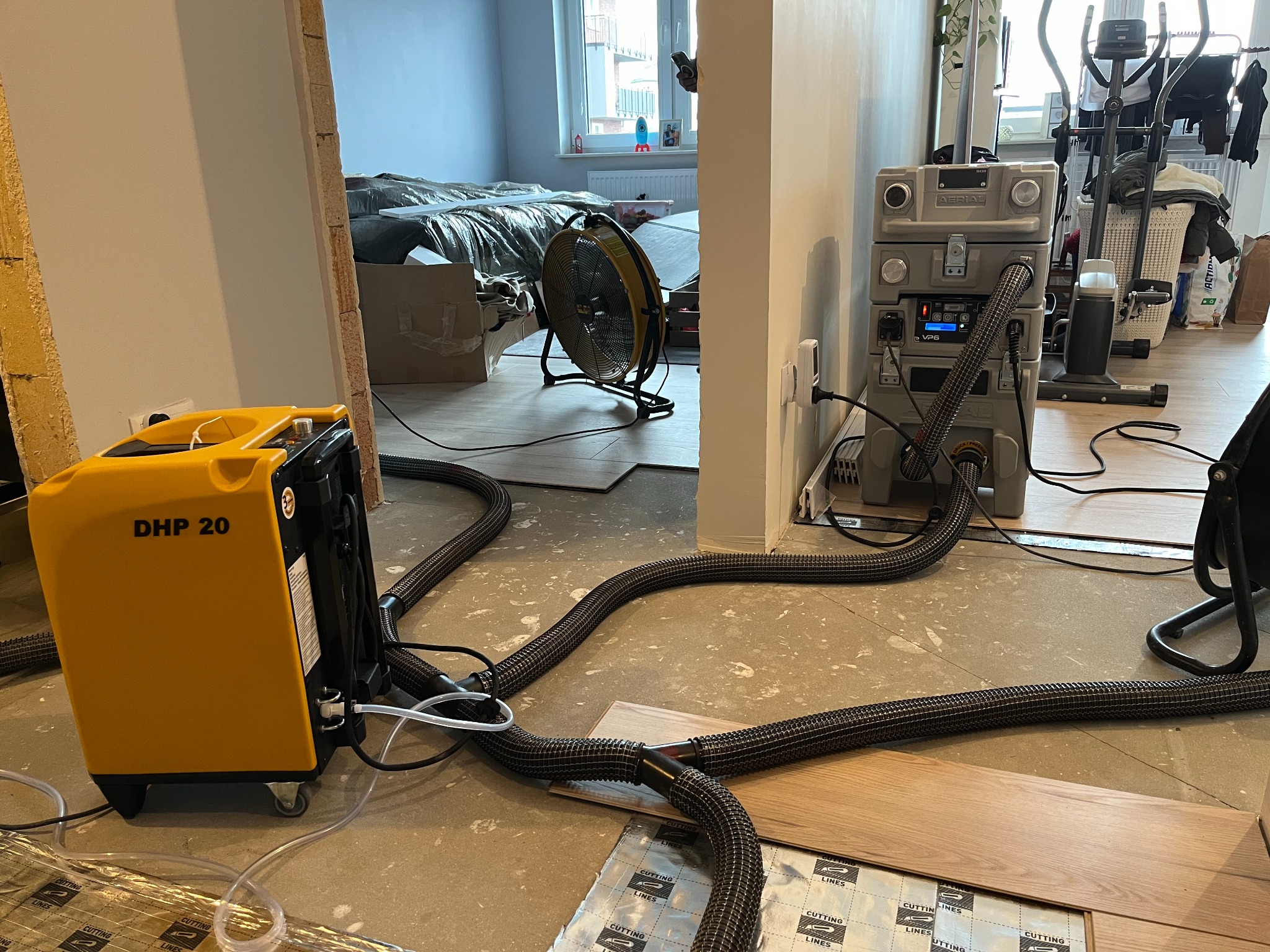 Żółty osuszacz DHP 20 w trakcie pracy, z podłączonymi wężami odprowadzającymi wilgoć, na tle przygotowanego podłoża pod parkiet i sprzętu do ćwiczeń w tle.