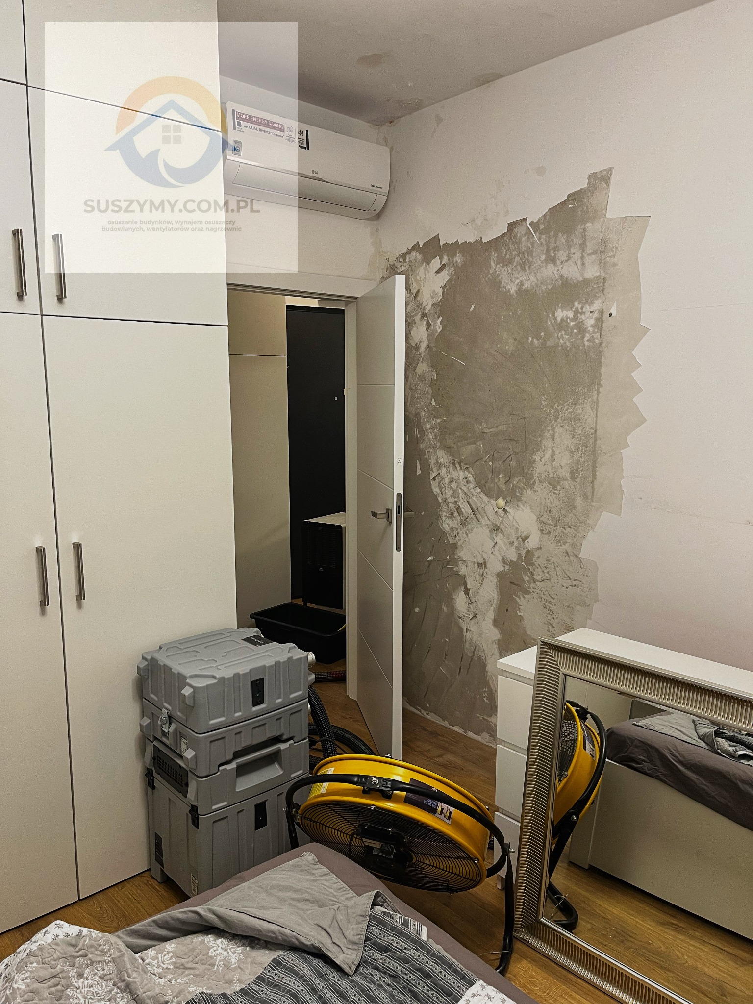 Pokój po zalaniu z uszkodzoną ścianą, widoczny osuszacz powietrza, wentylator i sprzęt w szarych skrzyniach. Na ścianie widoczne ślady po odpadającej farbie i wilgoci. W tle klimatyzator LG i logo...