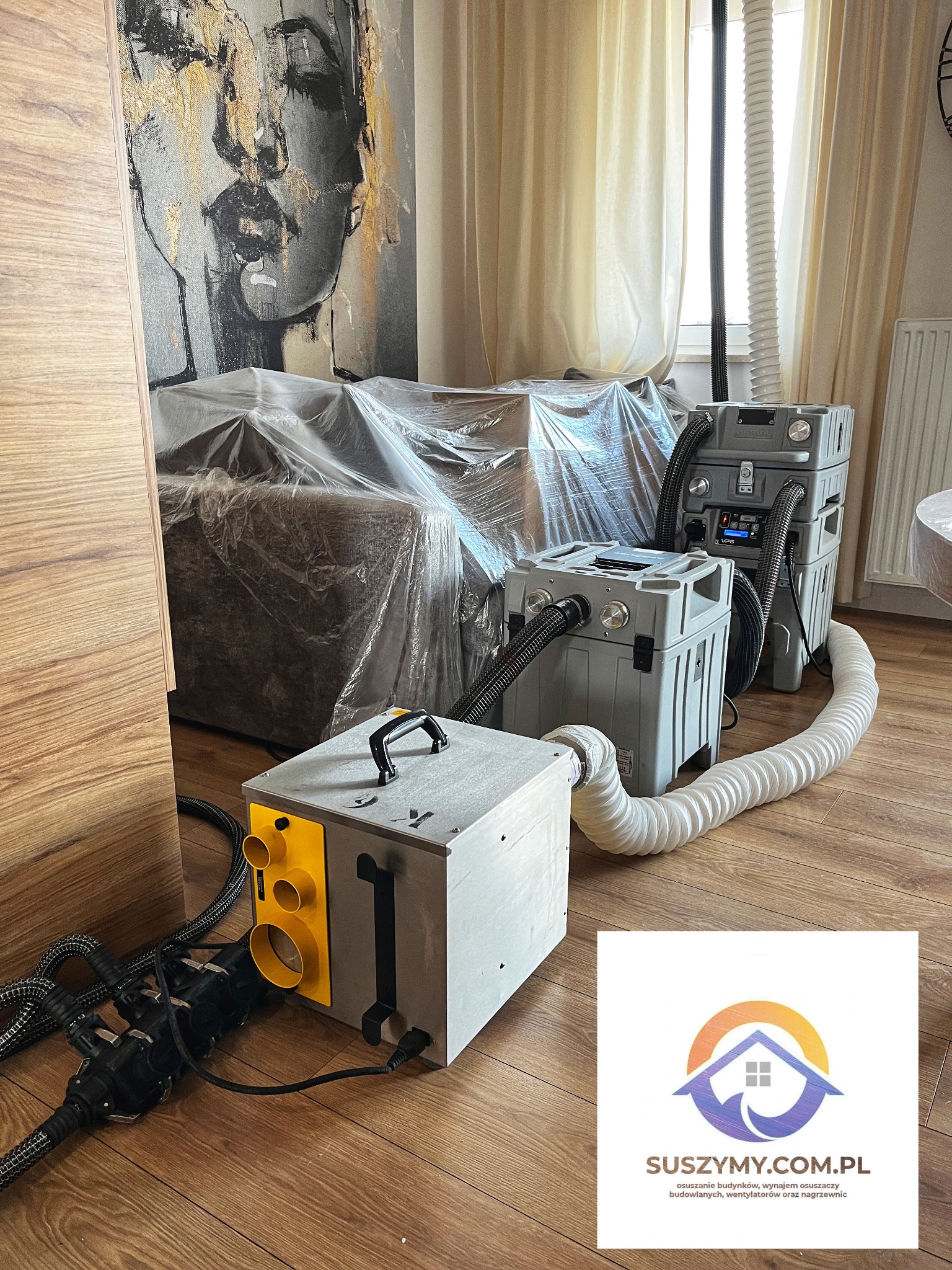 Wnętrze pokoju z meblami zabezpieczonymi folią i ustawionymi osuszaczami powietrza z widocznymi przewodami wentylacyjnymi; na podłodze widoczne logo firmy suszymy.com.pl