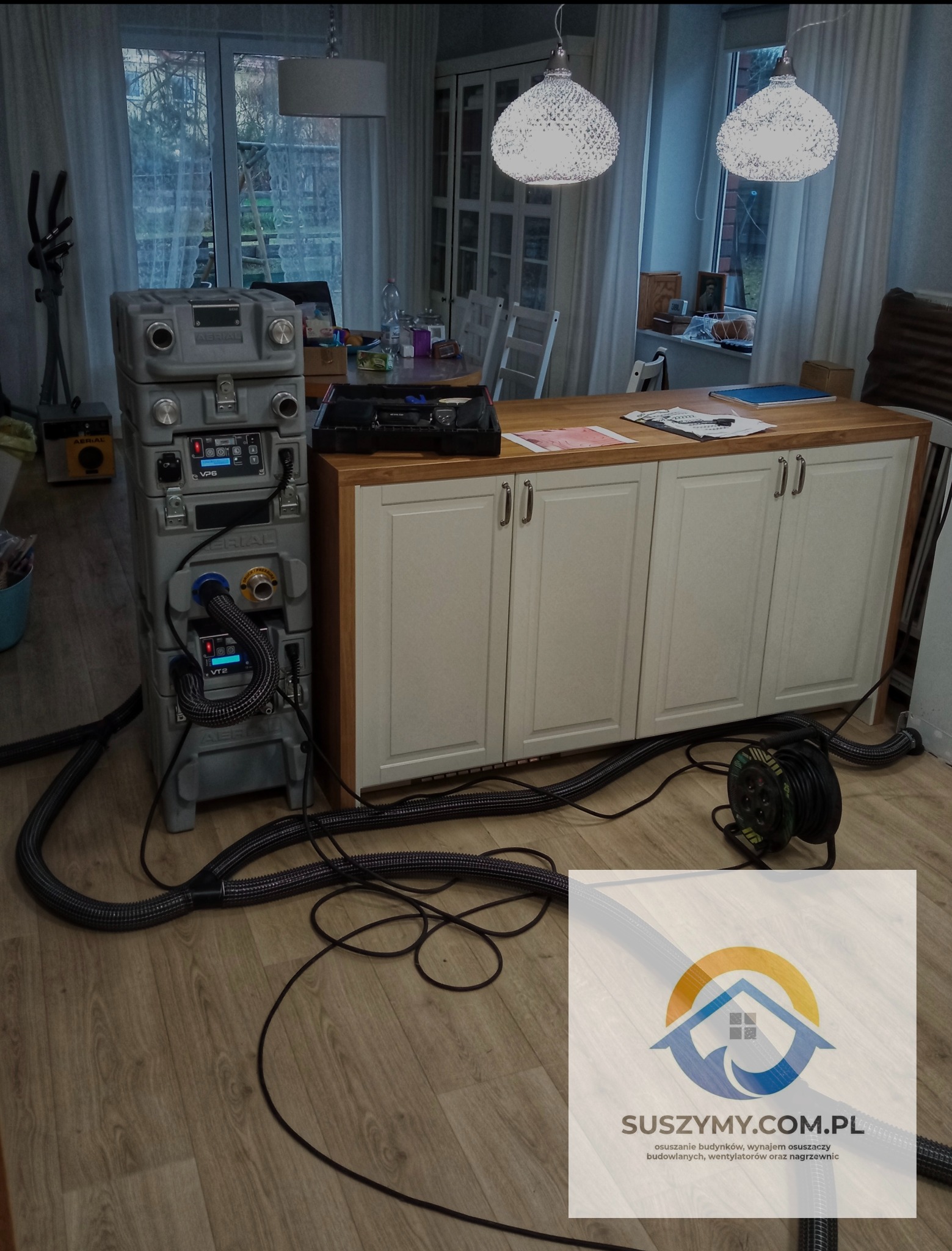 Profesjonalny osuszacz powietrza AERIAL VP6 i VT2 ustawiony w pomieszczeniu z drewnianą podłogą i białymi meblami, widoczne węże i przewody, w tle stół i krzesła, na pierwszym planie częściowo...