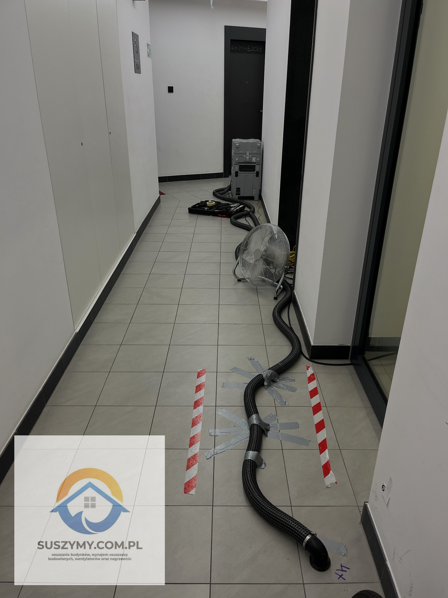 Korytarz w trakcie osuszania: widoczny wentylator, elastyczne rury przymocowane taśmą do podłogi wyznaczonej taśmą ostrzegawczą, w tle skrzynki narzędziowe, na podłodze logo firmy osuszającej.