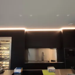 Nowoczesna kuchnia z czarnymi szafkami, oświetleniem LED podszafkowym i sufitowym, oraz wbudowaną chłodziarką na wino. Widoczny fragment blatu z pudełkami.