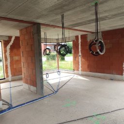 WatVolt INSTAL - Wnętrze w surowym stanie podczas prac elektrycznych: widoczne przewody poprowadzone po podłodze i zwisające z sufitu, ściany z czerwonej cegły, okna z widokiem na zieleń na zewnątrz.