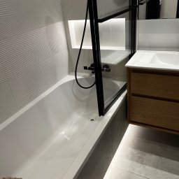 WatVolt INSTAL - Nowoczesna łazienka z białą wanną, kabiną prysznicową z czarnymi profilami, drewnianą szafką pod umywalką i geometrycznymi płytkami na ścianie.