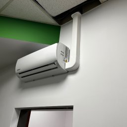 WatVolt INSTAL - Zamontowana biała klimatyzacja Kaisai na białej ścianie, z widocznymi rurami odprowadzającymi do sufitu. Fragment zielonego elementu architektonicznego w górnym rogu.