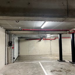 WatVolt INSTAL - Wnętrze garażu podziemnego z betonowymi ścianami i sufitem, widoczne instalacje elektryczne w rurach, oświetlenie fluorescencyjne, czerwone rury przeciwpożarowe, brama garażowa z automatem i dwie...