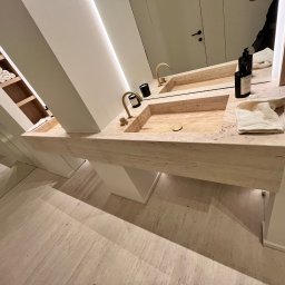 WatVolt INSTAL - Nowoczesna łazienka z podwójną umywalką z trawertynu, złotymi bateriami, minimalistycznymi dozownikami mydła i ręcznikami w kremowym odcieniu, oświetlona listwą LED pod lustrem.