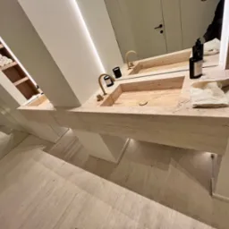 Nowoczesna łazienka z podwójną umywalką z trawertynu, złotymi bateriami, minimalistycznymi dozownikami mydła i ręcznikami w kremowym odcieniu, oświetlona listwą LED pod lustrem.