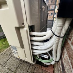 WatVolt INSTAL - Zewnętrzna jednostka klimatyzacji z podłączonymi przewodami chłodniczymi i elektrycznymi, zamocowanymi do ceglanej ściany budynku, widok z dołu.