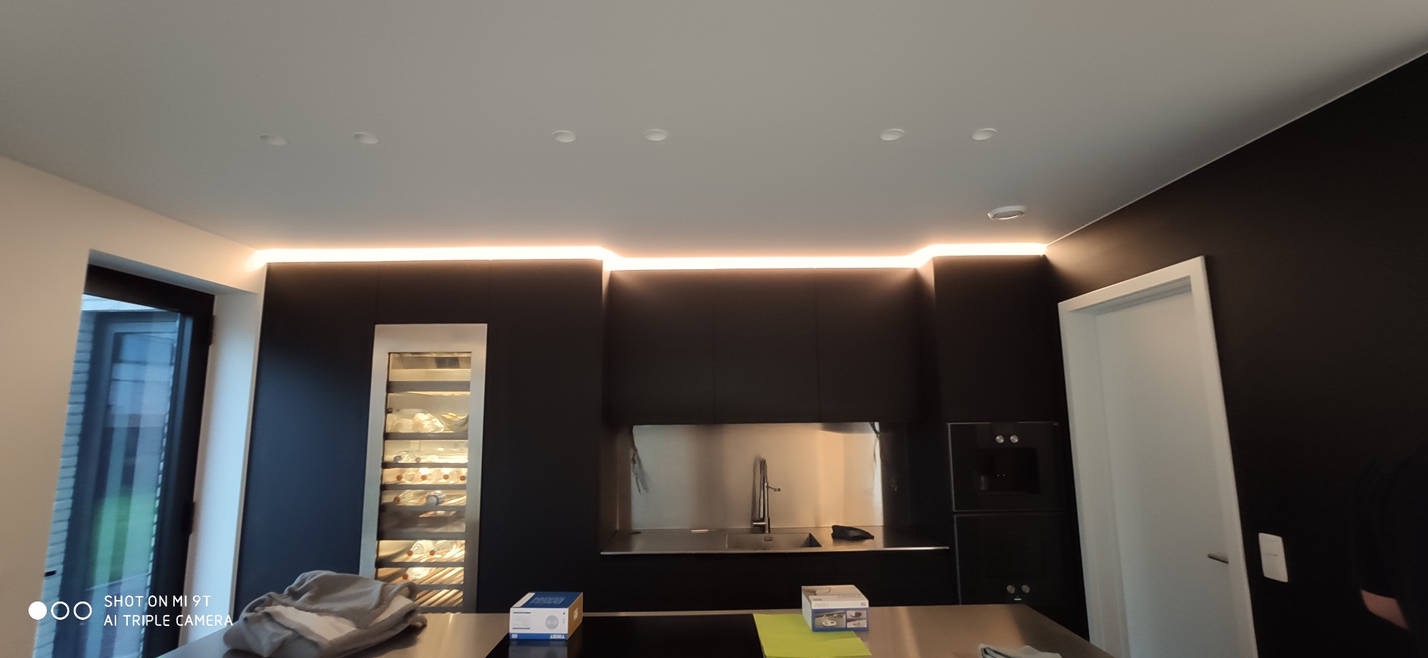 Nowoczesna kuchnia z czarnymi szafkami, oświetleniem LED podszafkowym i sufitowym, oraz wbudowaną chłodziarką na wino. Widoczny fragment blatu z pudełkami.