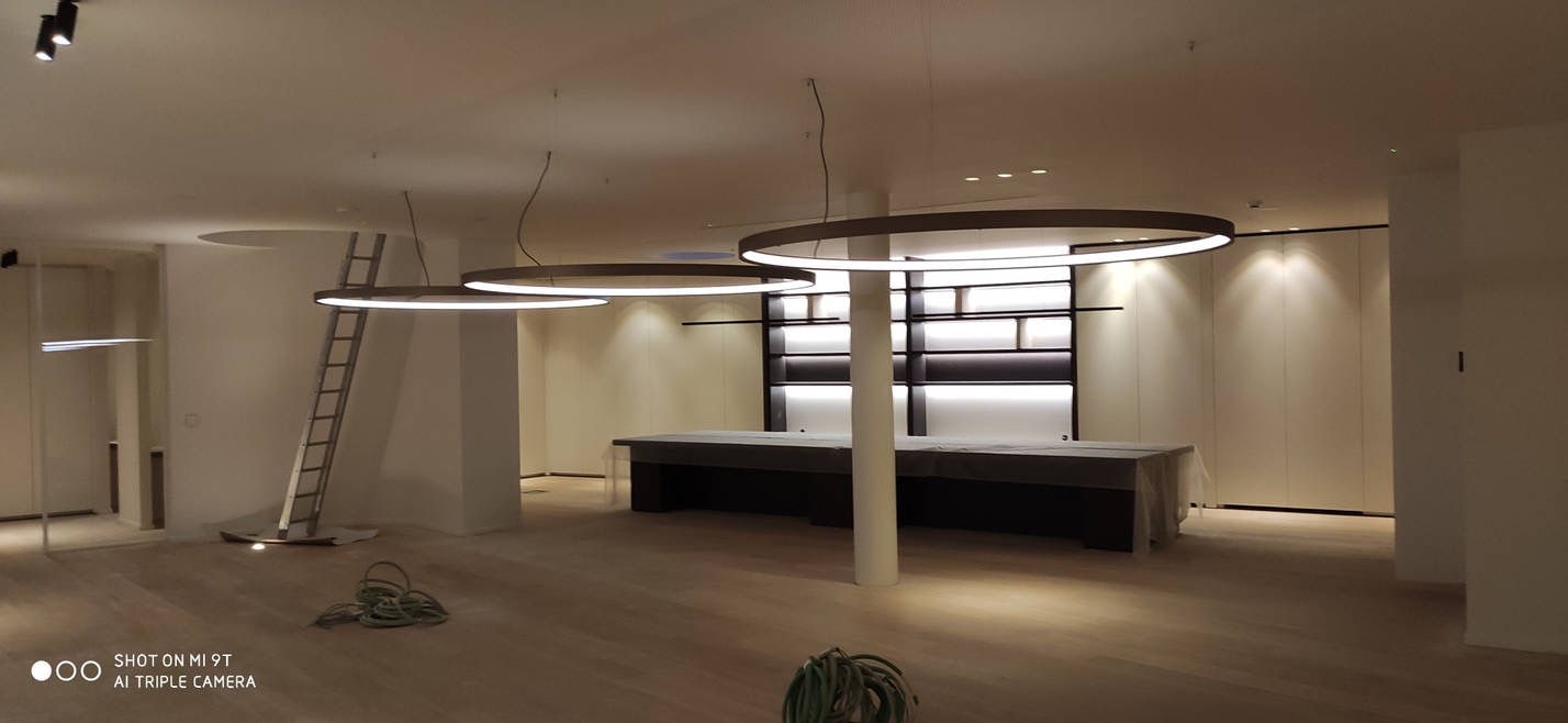 Wnętrze sklepu w trakcie instalacji oświetlenia: trzy okrągłe lampy LED zawieszone pod sufitem, drabina oparta o filar, przewody na podłodze, meble zakryte folią ochronną.