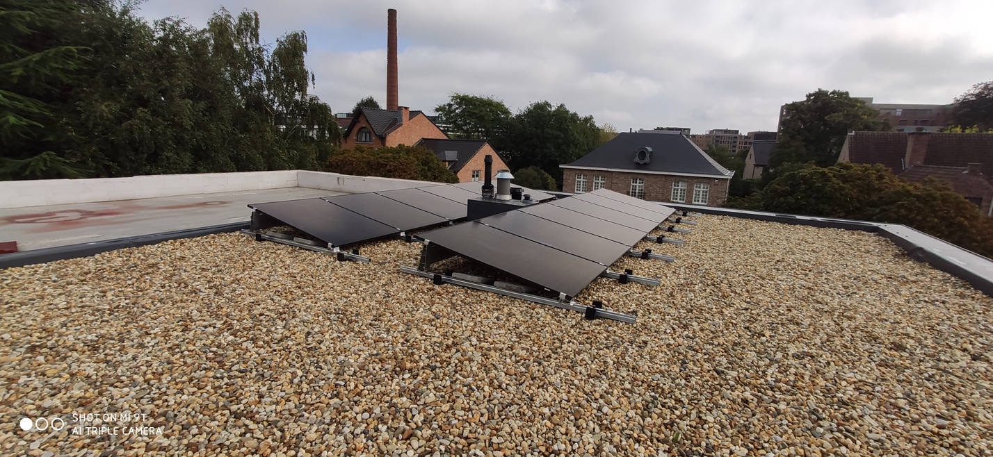 Panele słoneczne zamontowane na płaskim dachu pokrytym drobnym żwirem, w tle ceglane budynki i komin.