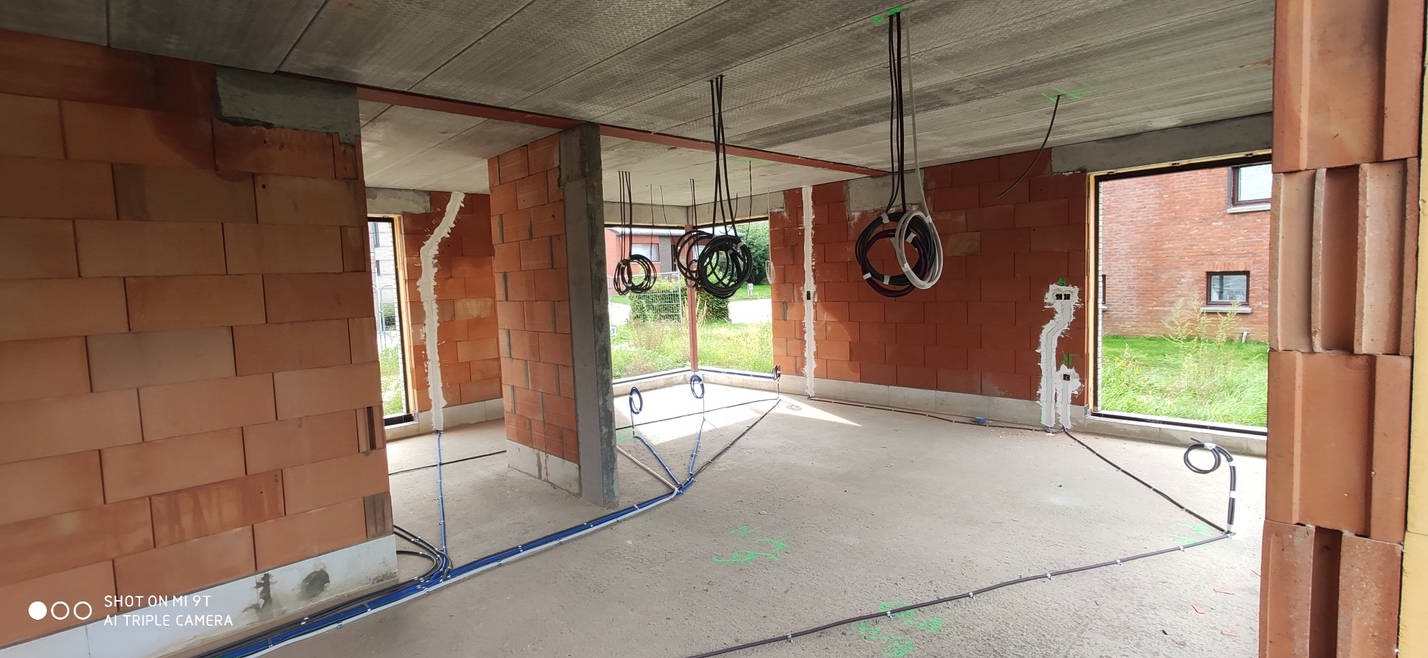 Wnętrze w surowym stanie podczas prac elektrycznych: widoczne przewody poprowadzone po podłodze i zwisające z sufitu, ściany z czerwonej cegły, okna z widokiem na zieleń na zewnątrz.