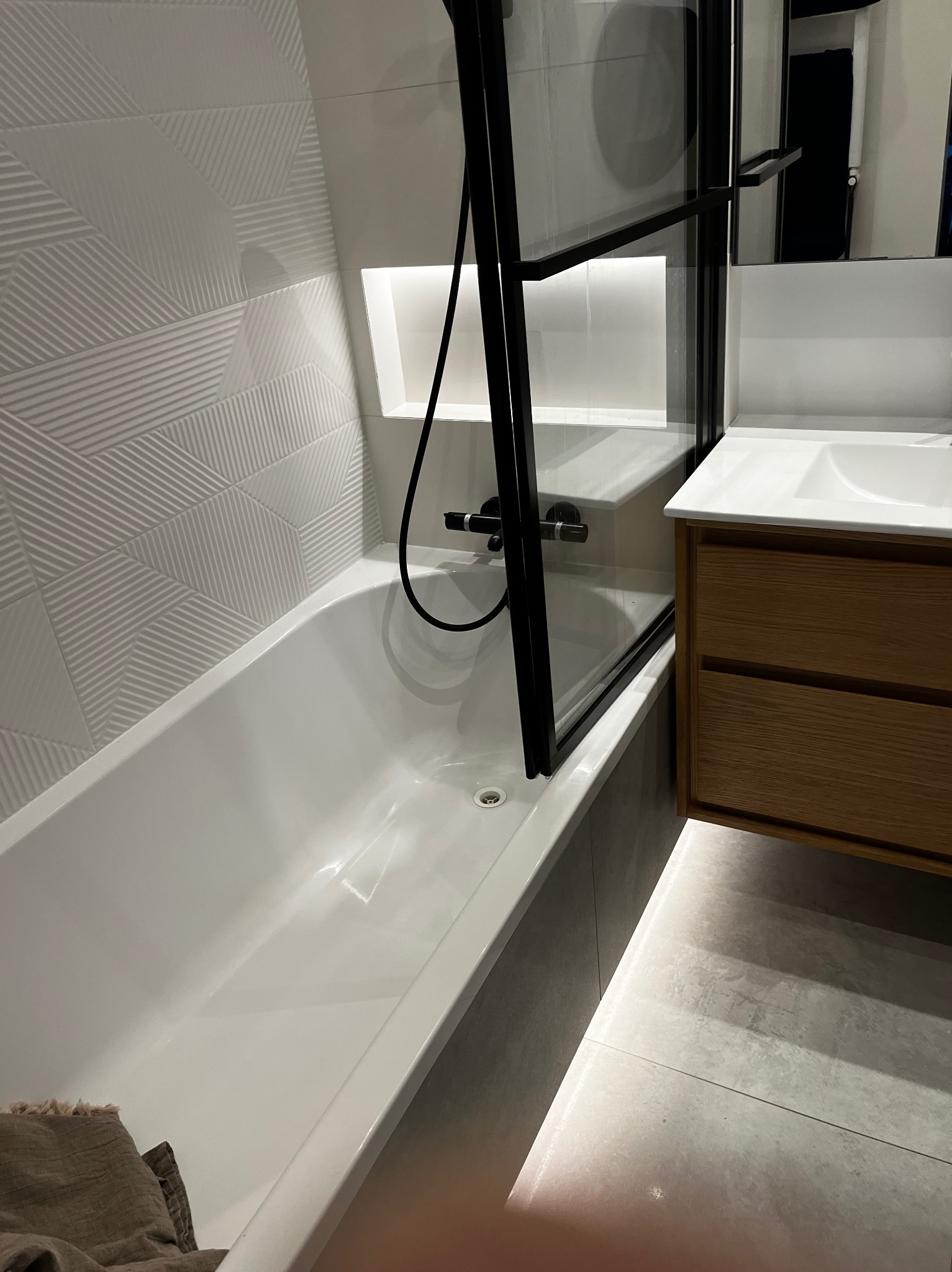 Nowoczesna łazienka z białą wanną, kabiną prysznicową z czarnymi profilami, drewnianą szafką pod umywalką i geometrycznymi płytkami na ścianie.