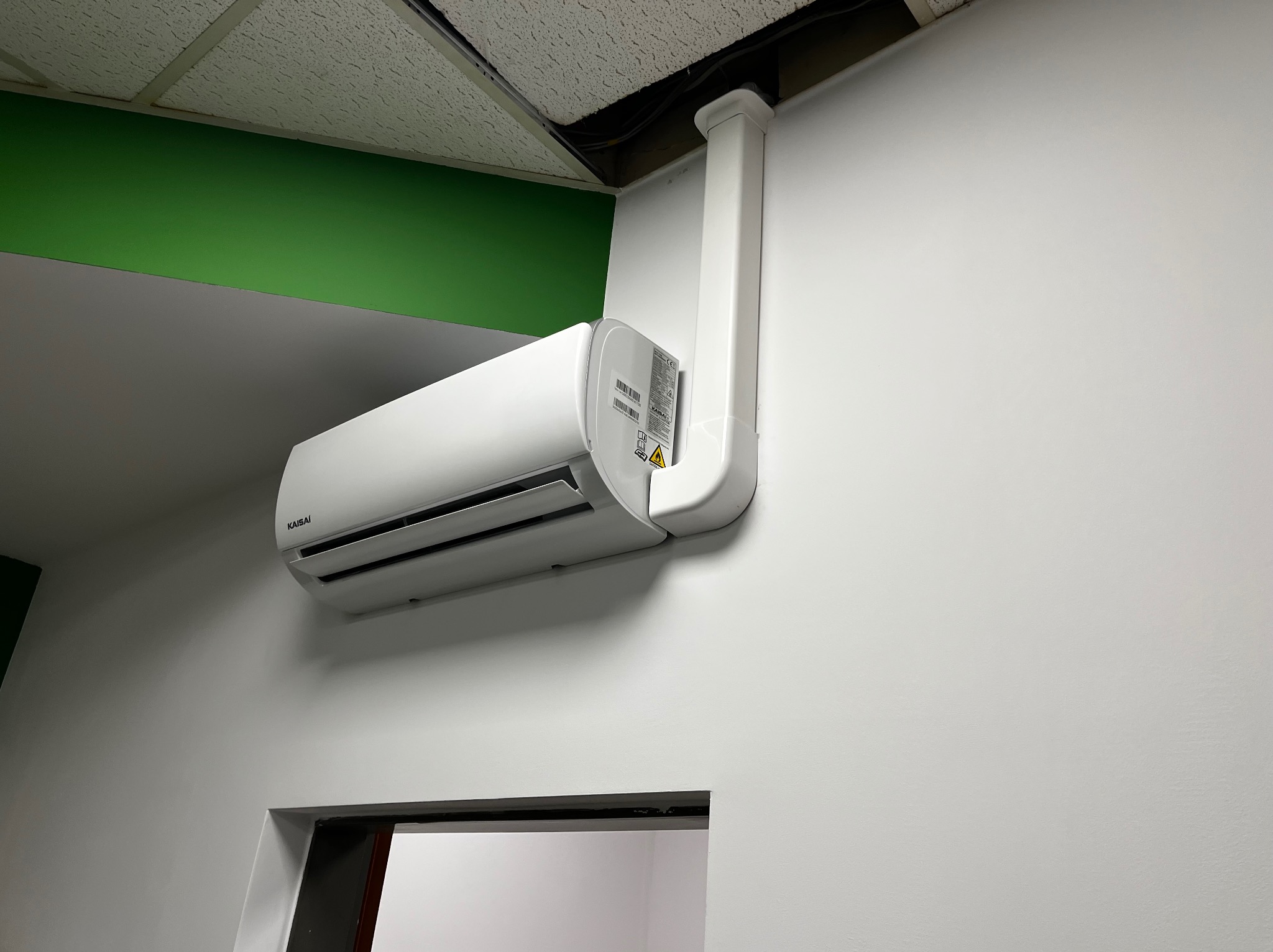 Zamontowana biała klimatyzacja Kaisai na białej ścianie, z widocznymi rurami odprowadzającymi do sufitu. Fragment zielonego elementu architektonicznego w górnym rogu.