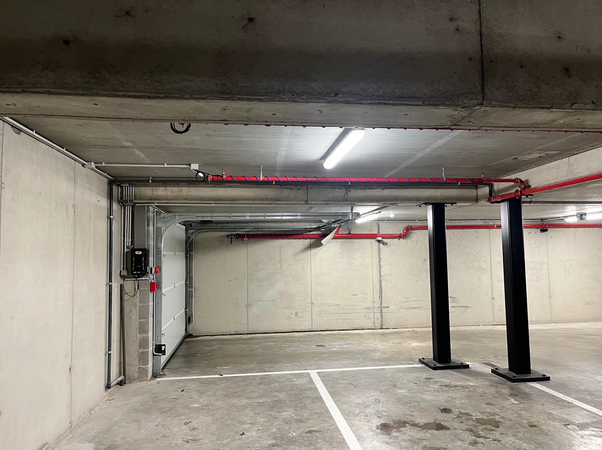 Wnętrze garażu podziemnego z betonowymi ścianami i sufitem, widoczne instalacje elektryczne w rurach, oświetlenie fluorescencyjne, czerwone rury przeciwpożarowe, brama garażowa z automatem i dwie...