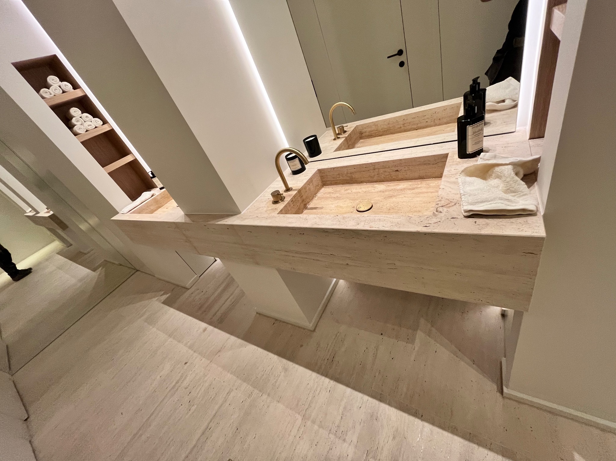Nowoczesna łazienka z podwójną umywalką z trawertynu, złotymi bateriami, minimalistycznymi dozownikami mydła i ręcznikami w kremowym odcieniu, oświetlona listwą LED pod lustrem.