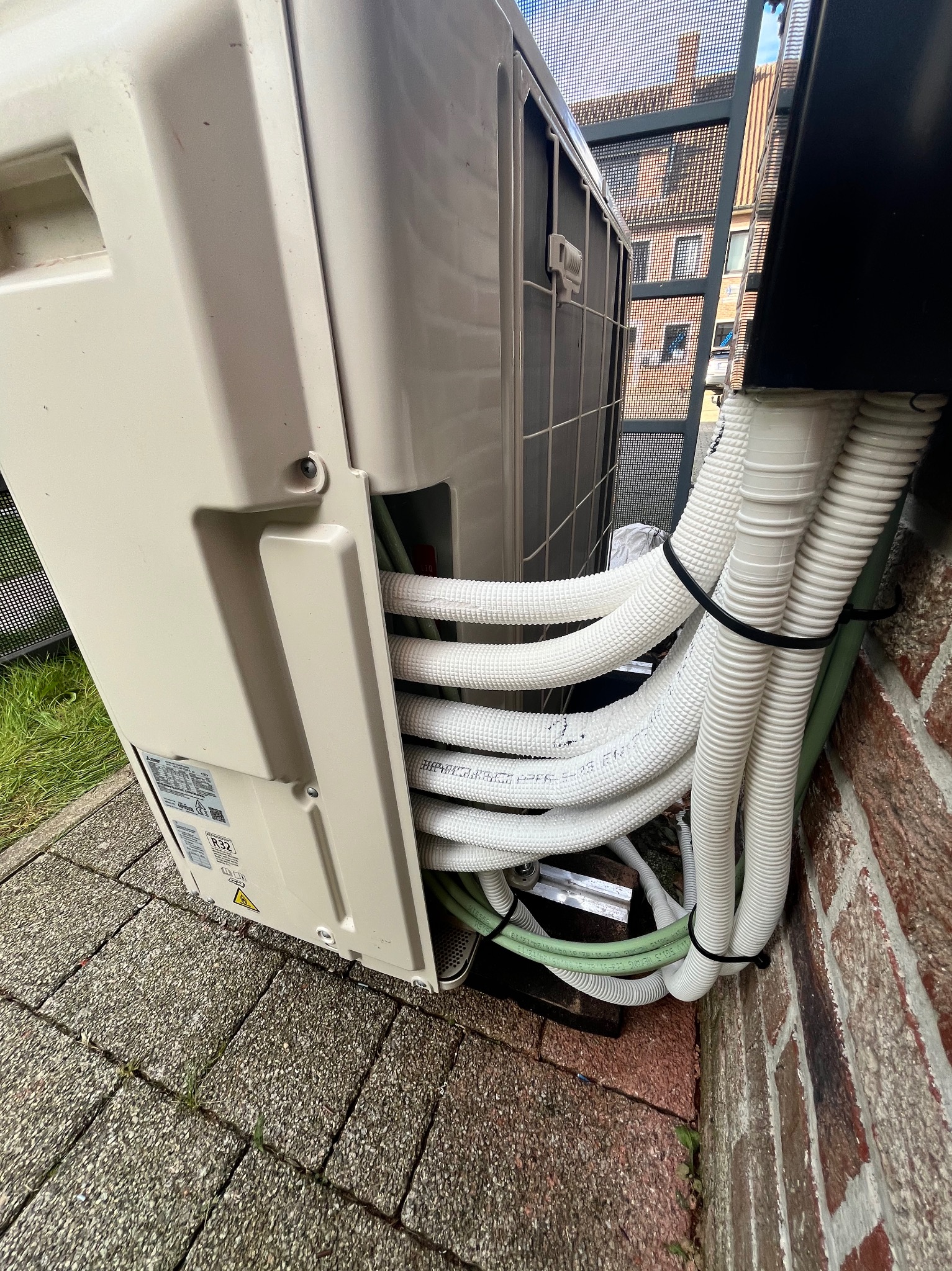 Zewnętrzna jednostka klimatyzacji z podłączonymi przewodami chłodniczymi i elektrycznymi, zamocowanymi do ceglanej ściany budynku, widok z dołu.