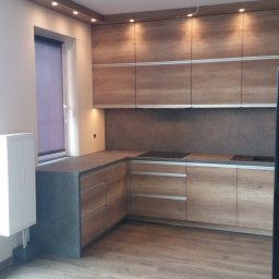 Tomasz Krajęta - Nowoczesna kuchnia z drewnianymi frontami szafek, szarym blatem i podłogą z paneli imitujących drewno. Górne szafki podświetlone punktowymi lampami.