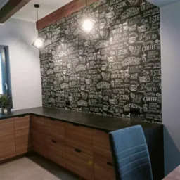 Kuchnia z drewnianymi szafkami i ścianą pokrytą tablicową tapetą z rysunkami kawy, steków i pieczywa, oświetlona dwoma wiszącymi lampami.