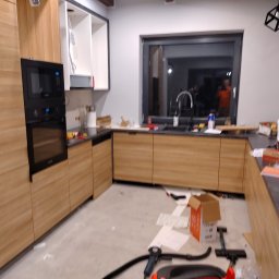 Tomasz Krajęta - Kuchnia w trakcie montażu: widoczne szafki z jasnego drewna, czarny piekarnik w zabudowie, czarne blaty, okno z czarną ramą, na podłodze kartony, papiery, narzędzia i odkurzacz przemysłowy.