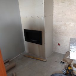 Tomasz Krajęta - Wnętrze w trakcie wykończenia: obudowa kominka z płyt kartonowo-gipsowych z wnęką, fragment ściany wyłożonej dekoracyjnym kamieniem, narzędzia budowlane na podłodze.