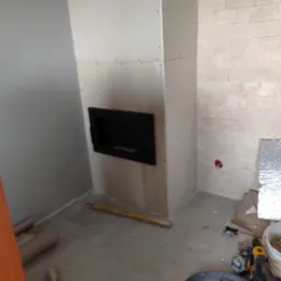 Wnętrze w trakcie wykończenia: obudowa kominka z płyt kartonowo-gipsowych z wnęką, fragment ściany wyłożonej dekoracyjnym kamieniem, narzędzia budowlane na podłodze.