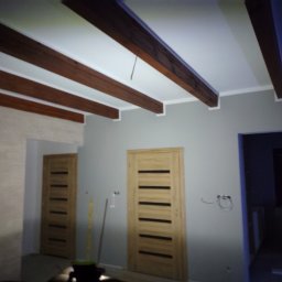 Tomasz Krajęta - Wnętrze pokoju z drewnianymi belkami sufitowymi, szarą ścianą, dwiema drewnianymi ościeżnicami z zamontowanymi drzwiami i widocznymi przewodami elektrycznymi wychodzącymi ze ściany.