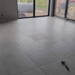 Szare płytki podłogowe ułożone w nowym domu, widok przez okno na zewnątrz, gdzie stoi czerwony traktor, młotek leży na podłodze.