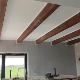 Tomasz Krajęta - Wnętrze w trakcie wykańczania: pomalowany sufit z drewnianymi belkami, szara ściana, duże okno z widokiem na zielone pola, oraz rusztowanie z narzędziami i siedzący mężczyzna