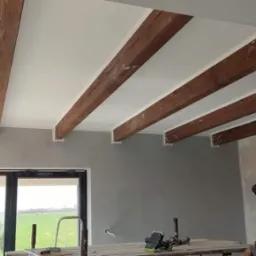 Wnętrze w trakcie wykańczania: pomalowany sufit z drewnianymi belkami, szara ściana, duże okno z widokiem na zielone pola, oraz rusztowanie z narzędziami i siedzący mężczyzna