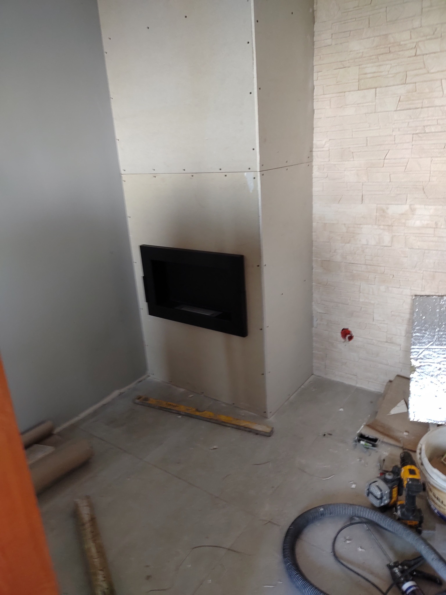 Wnętrze w trakcie wykończenia: obudowa kominka z płyt kartonowo-gipsowych z wnęką, fragment ściany wyłożonej dekoracyjnym kamieniem, narzędzia budowlane na podłodze.