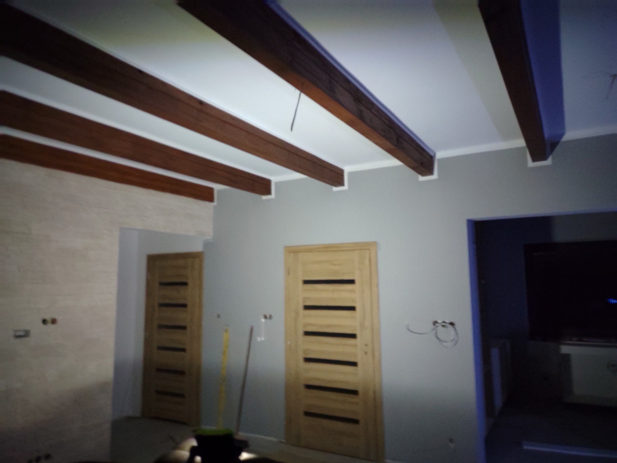 Wnętrze pokoju z drewnianymi belkami sufitowymi, szarą ścianą, dwiema drewnianymi ościeżnicami z zamontowanymi drzwiami i widocznymi przewodami elektrycznymi wychodzącymi ze ściany.