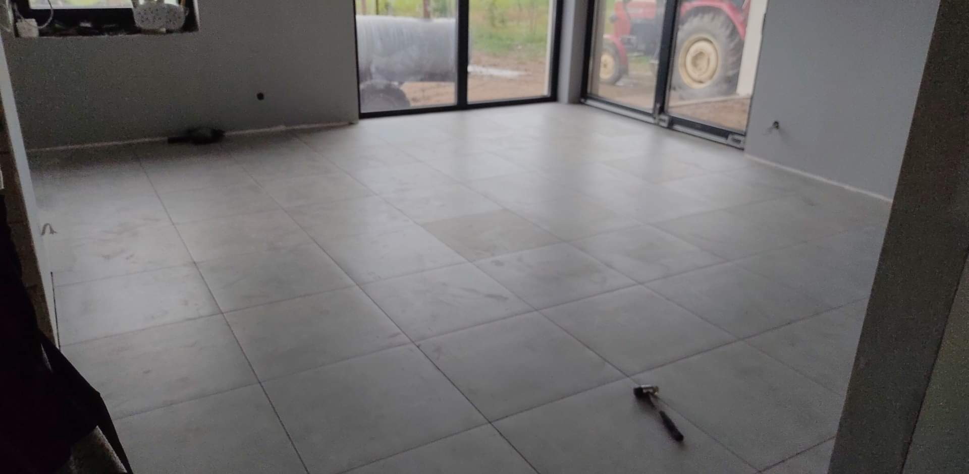 Szare płytki podłogowe ułożone w nowym domu, widok przez okno na zewnątrz, gdzie stoi czerwony traktor, młotek leży na podłodze.