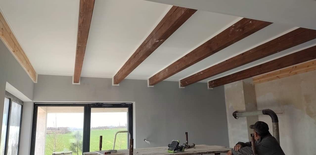 Wnętrze w trakcie wykańczania: pomalowany sufit z drewnianymi belkami, szara ściana, duże okno z widokiem na zielone pola, oraz rusztowanie z narzędziami i siedzący mężczyzna