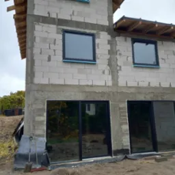 Budynek w trakcie budowy z zamontowanymi oknami i drzwiami przesuwnymi w czarnych ramach, widoczne narzędzia budowlane i przygotowany teren.