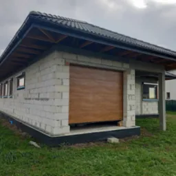 Surowy budynek garażu z brązową segmentową bramą, częściowo otwartą, widoczne bloczki betonowe i elementy konstrukcji dachu, szara dachówka, trawa przed budynkiem.