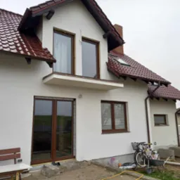 Biały dom jednorodzinny z brązowymi ramami okiennymi i drzwiami przesuwnymi, widoczne elementy montażowe i rower zaparkowany obok ściany.