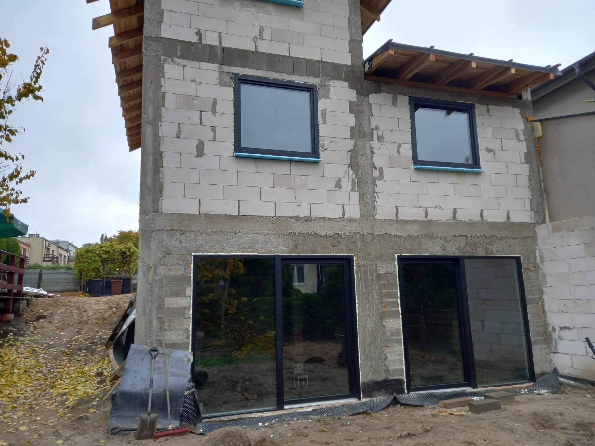 Budynek w trakcie budowy z zamontowanymi oknami i drzwiami przesuwnymi w czarnych ramach, widoczne narzędzia budowlane i przygotowany teren.