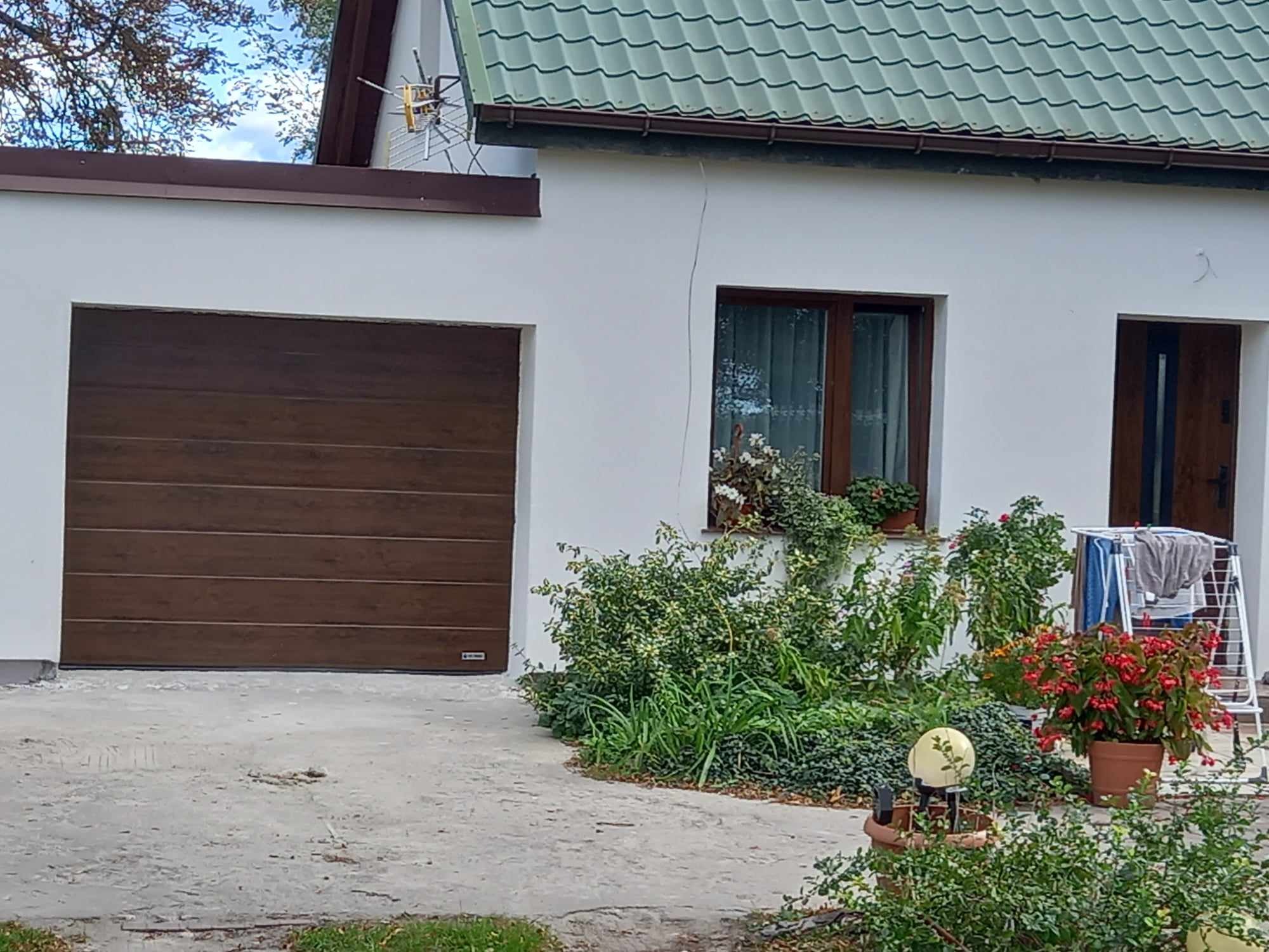 Brązowa brama garażowa segmentowa, z poziomymi panelami, w białym budynku z zielonym dachem, widok z zewnątrz, z elementami ogrodu i suszarką na pranie.