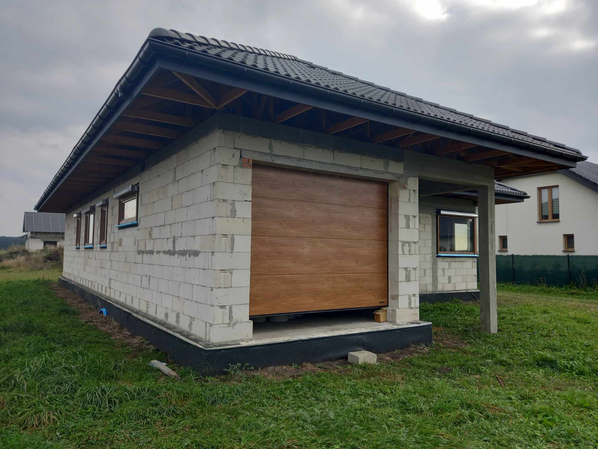 Surowy budynek garażu z brązową segmentową bramą, częściowo otwartą, widoczne bloczki betonowe i elementy konstrukcji dachu, szara dachówka, trawa przed budynkiem.