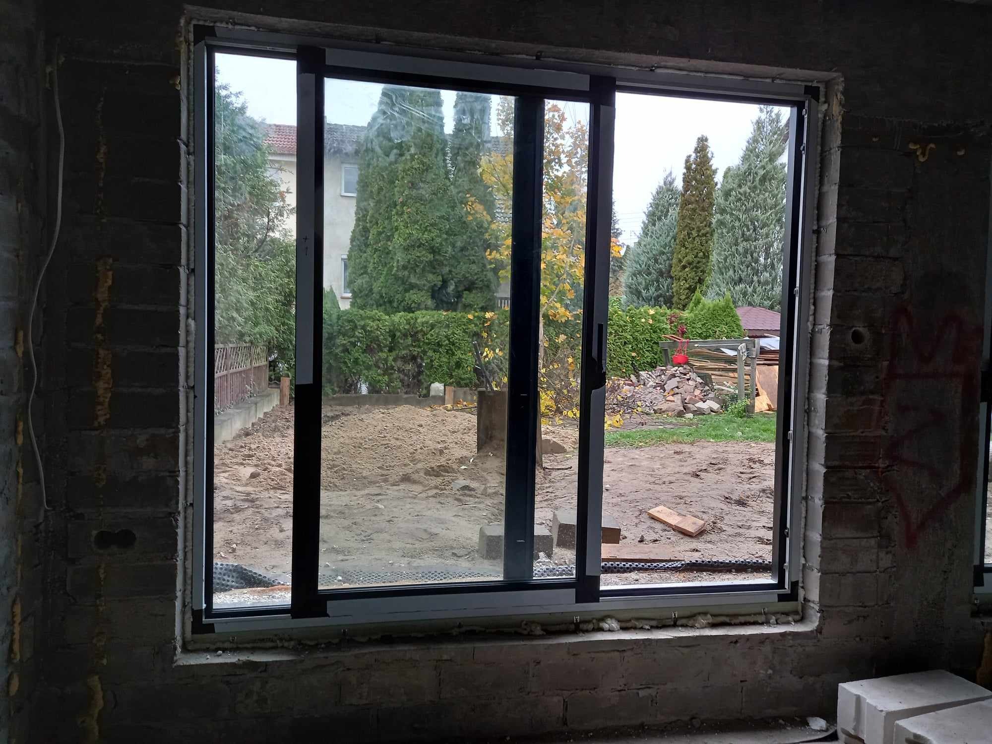 Trzyczęściowe okno przesuwne z czarną ramą, widok z surowego wnętrza na ogród w Bydgoszczy, częściowo otwarte skrzydło.