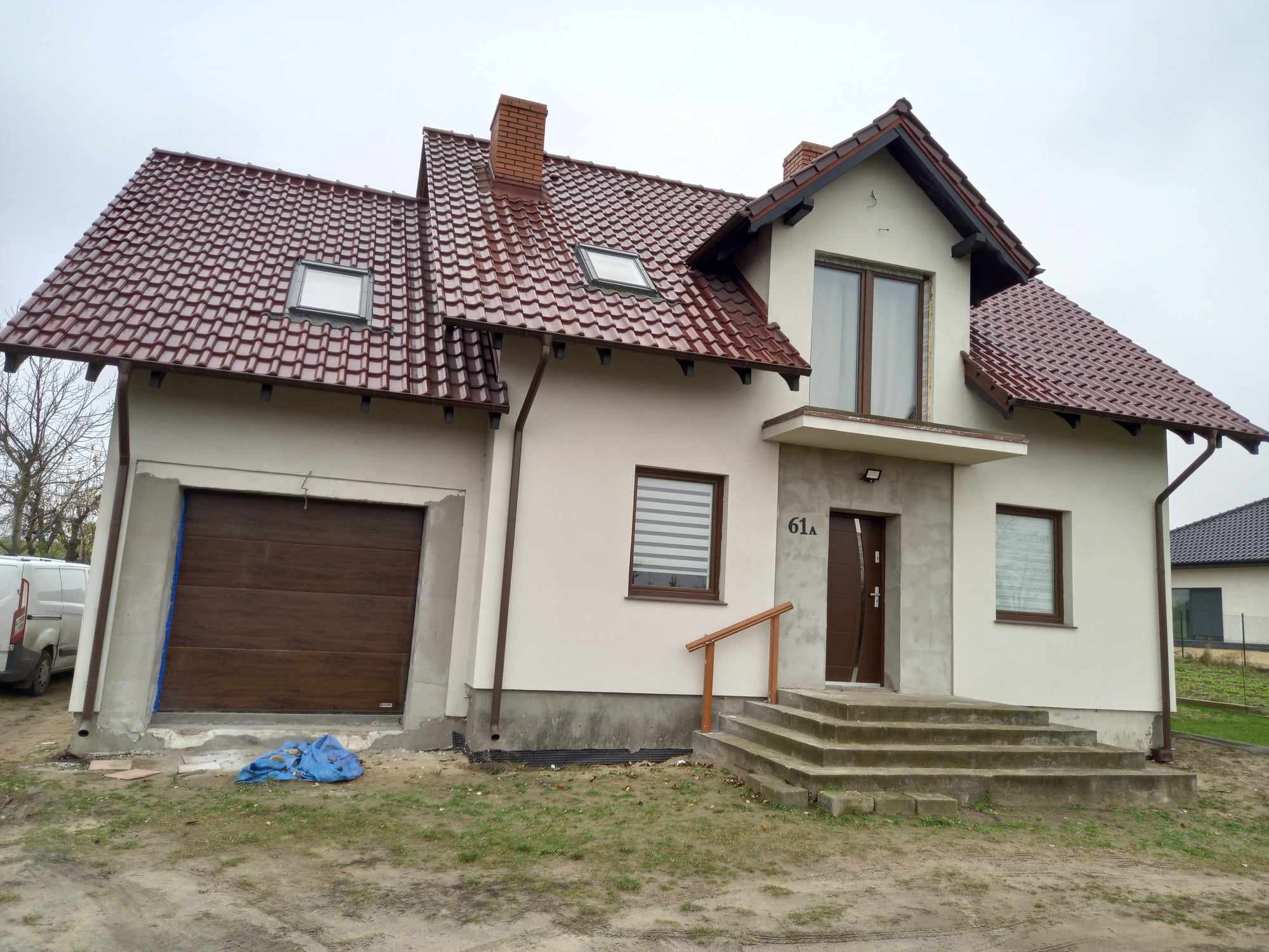 Nowo wybudowany dom z brązową stolarką okienną i drzwiową, częściowo wykończony, z widocznymi elementami podłączeń i zabezpieczeniami budowlanymi.